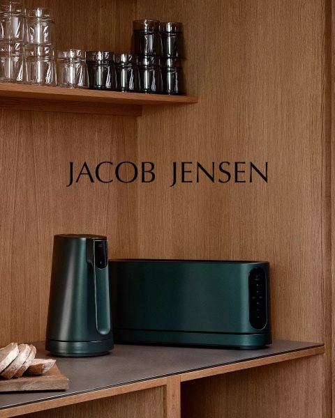 Una cocina moderna con estantes de madera, vasos, una hogaza de pan y electrodomésticos Jacob Jensen de color verde oscuro en la encimera. El nombre de la marca "JACOB JENSEN" está en la pared del panel de madera.