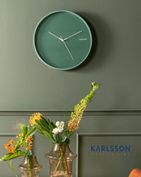 Un reloj de pared minimalista verde con manecillas blancas cuelga de una pared verde sobre dos jarrones de vidrio con diferentes flores naranjas, blancas y amarillas. En la esquina inferior derecha aparece en azul la palabra "KARLSSON".