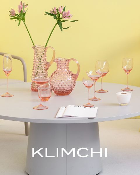 Una mesa elegantemente dispuesta presenta cristalería rosa, que incluye jarras, vasos y copas. Flores rosas en un jarrón añaden un toque de elegancia. El fondo es de un suave color amarillo y la palabra "KLIMCHI" aparece en la parte inferior de la imagen.