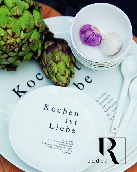 Se exhiben platos blancos con el texto en alemán "Cocinar es amor" junto con alcachofas y cuencos con ajo. El arreglo incluye cucharas blancas y el logo "Räder" en un plato.