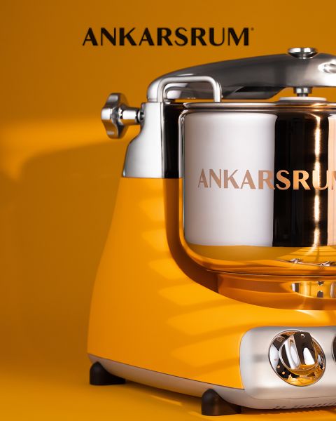 Primer plano de una batidora de cocina Ankarsrum amarilla con un brillante cuenco de acero inoxidable frente a un fondo amarillo a juego. El nombre de la marca Ankarsrum aparece encima de la máquina.