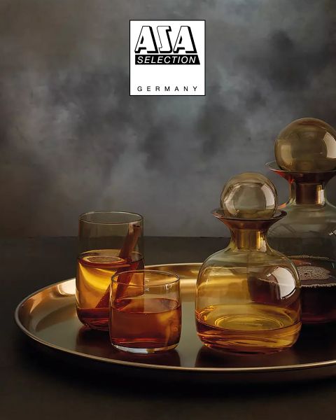 Jarras color ámbar con vasos a juego, llenas de un líquido marrón, se colocan sobre una bandeja dorada frente a un fondo texturizado gris atmosférico. En la parte superior se muestra el logotipo de ASA Selection Alemania.