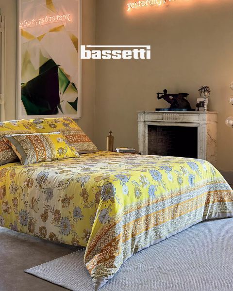 Un dormitorio con una colcha de flores amarillas, almohadas a juego, una chimenea de mármol blanco, arte moderno en la pared y un letrero de neón encima que dice "Ayer, hoy". La palabra "Bassetti" es claramente visible.