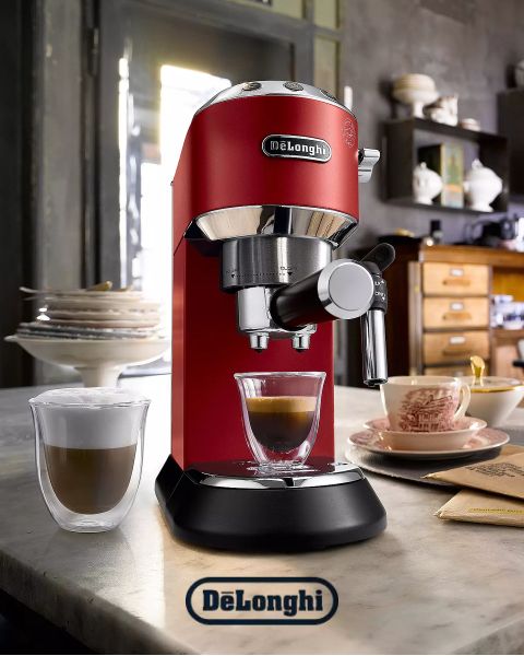 Una máquina de espresso De'Longhi roja en una encimera de cocina vierte café en una taza de vidrio. En el fondo se ve una taza de capuchino, una pila de panqueques y una cocina acogedora.