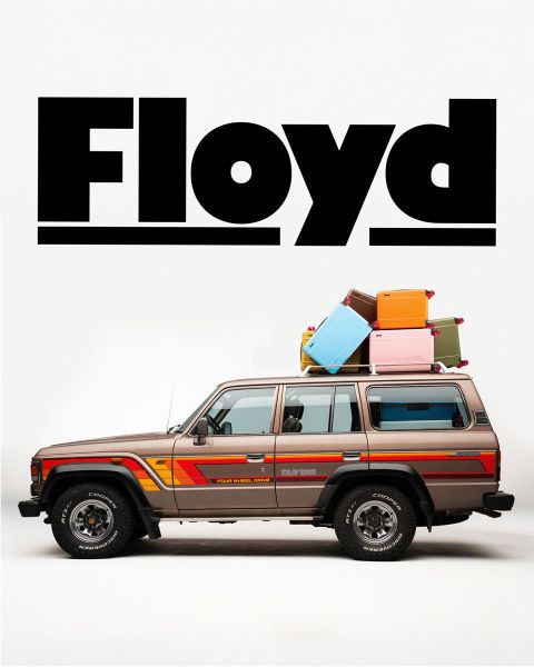 Un SUV clásico de diseño retro con equipaje en el techo está estacionado frente a un fondo blanco. Encima del vehículo, la palabra "Floyd" destaca en negritas letras negras.
