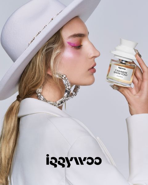 Una persona con pelo largo, un sombrero blanco y maquillaje de ojos rosa sostiene una botella que dice "Cashmere Show Pony" de Iggywoo. Lleva grandes aros brillantes y una chaqueta blanca delante de un fondo sencillo. Abajo se ve el logo de Iggywoo.