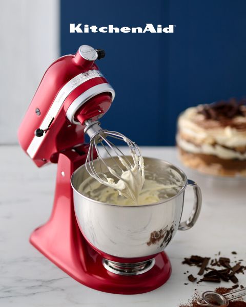 Una batidora KitchenAid roja con crema batida en el batidor de varillas sobre una placa de mármol. En el fondo se ven un postre en capas y virutas de chocolate, encima pone "KitchenAid".