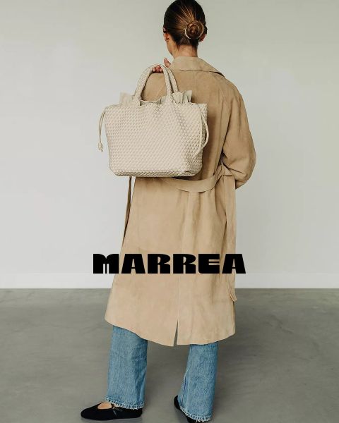 Una mujer con un moño pulcro, un abrigo largo beige, vaqueros azules y zapatos negros está de pie de espaldas sosteniendo un bolso grande tejido de color crema. En la parte baja de su espalda está la palabra "MARREA".
