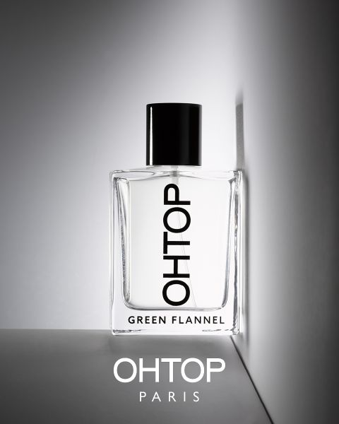 El frasco de perfume con la inscripción "OHTOP GREEN FLANNEL" se encuentra sobre una superficie gris delante de un fondo gris claro. La botella está parcialmente sombreada, debajo está la inscripción "OHTOP PARIS". Diseño minimalista con cierre negro.