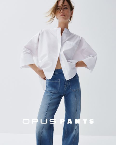 Persona con camisa blanca holgada y vaqueros azules, con las manos en las caderas. Abajo está el texto "Opus Pants". El cabello está ligeramente despeinado por el viento, fondo blanco.