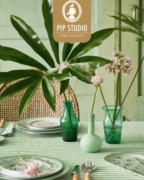 Sobre una mesa con un mantel de rayas verdes hay jarrones de cristal con ramas frondosas y flores rosas. Platos con diseños elaborados se colocan junto a los cubiertos. En el fondo se puede ver una silla de mimbre y un logotipo de Pip Studio.