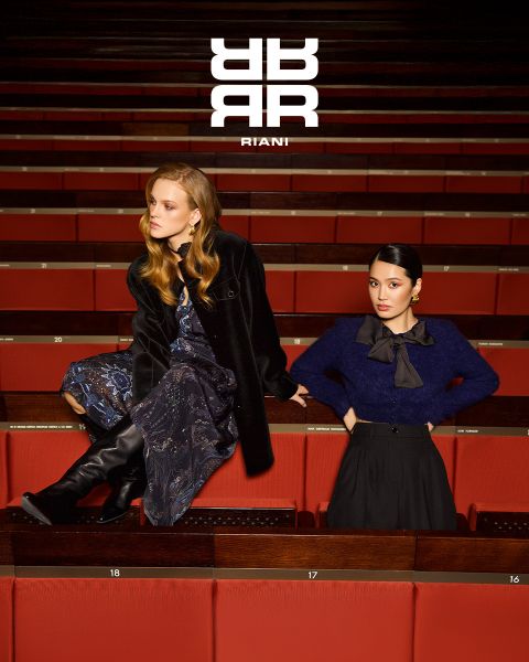 Dos mujeres posan con estilo en un teatro con asientos rojos. Una está sentada en el respaldo de un asiento, vestida con un vestido estampado, un abrigo negro y botas. La otra está de pie junto a ella con un jersey azul marino y una falda negra. Encima de ellas aparece el logo "RIANI".