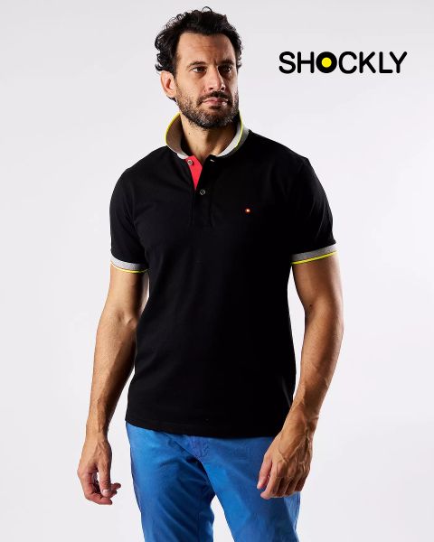 Un hombre con una camisa polo negra con detalles en amarillo y blanco y pantalones azules está de pie frente a un fondo blanco liso. En la parte superior derecha está la palabra "SHOCKLY" en letras negras.