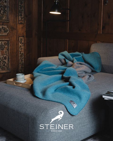 Un salón acogedor con un sofá gris, una manta azul, una bandeja de madera con taza y cuenco, una lámpara de pie y paredes revestidas de madera. Abajo se ve el logo de "STEINER".