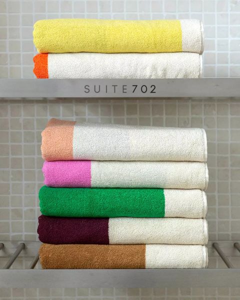 Seis toallas dobladas pulcramente con extremos de colores: amarillo, naranja, marrón, rosa, verde y burdeos, apiladas en dos estantes frente a una pared de azulejos. En el estante superior está escrito "SUITE702".