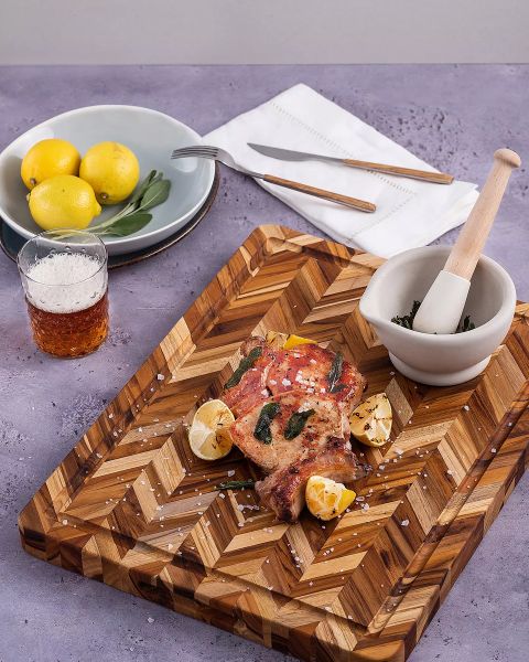 Una tabla de cortar de madera con carne cocida y especiada, gajos de limón y sal gruesa. Al lado hay un mortero con hierbas, un vaso con bebida, un plato con limones, así como un tenedor y un cuchillo sobre una servilleta, todo sobre una superficie violeta.