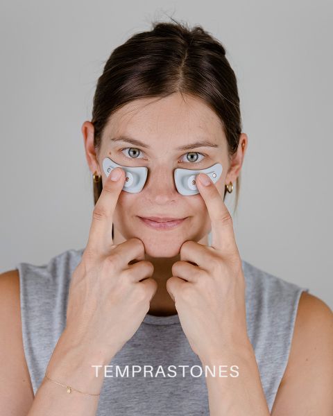 Una persona sostiene dos pequeños dispositivos ovalados debajo de los ojos y los presiona suavemente contra su piel. Tiene una expresión facial neutra y lleva una blusa sin mangas. En la parte inferior de la imagen aparece la palabra "TEMPRASTONES".