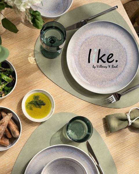 Vista superior de una mesa de comedor con un plato blanco y azul que dice "like. by Villeroy & Boch", manteles individuales verdes, vasos de vidrio transparente, un tazón de grissini, un tazón de ensalada y un tazón de aceite, adornado con hierbas.