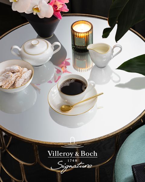 En una mesa de centro de cristal reflectante hay una taza de café en una taza blanca y dorada, una pequeña tetera, una jarrita de leche, una vela y un plato con galletas. La escena parece elegante y acogedora, y en la parte inferior se ve el logotipo de Villeroy & Boch.