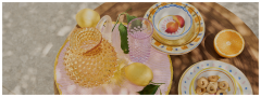 Una mesa de madera con una jarra de vidrio texturizado, un vaso rosa, limones enteros con hojas, platos decorativos apilados, media naranja y un plato con pequeñas galletas redondas bajo la luz del sol moteada.