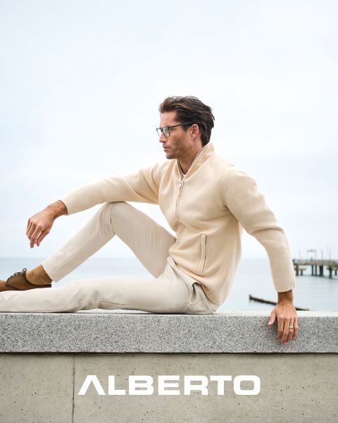 Un hombre con gafas, un jersey beige con cremallera y un pantalón claro está sentado en un saliente de piedra junto al mar. Mira hacia un lado, debajo de él se lee "ALBERTO" en letras blancas. El fondo está nublado y se ve un muelle.