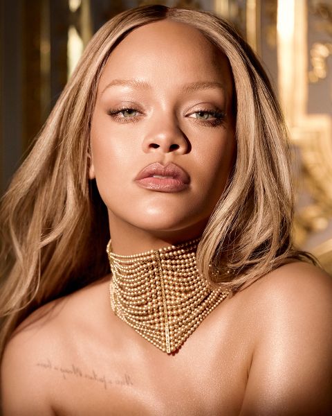 Una mujer con cabello rubio largo y liso y un collar de perlas doradas posa en un entorno suavemente iluminado con decoración dorada. Lleva maquillaje neutro y un tatuaje visible en la clavícula.