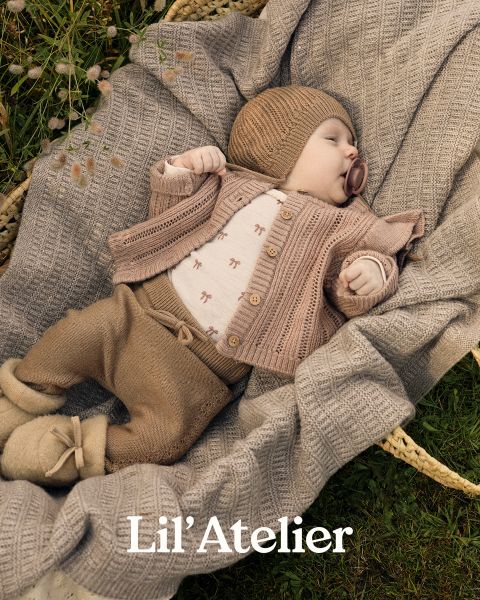 Un bebé con un gorro de punto marrón, una chaqueta de punto y pantalones está acostado sobre una manta beige texturizada en una canasta al aire libre. El bebé tiene un chupete y pies pequeños. El texto en la parte inferior dice "Lil' Atelier".