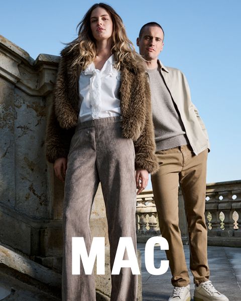 Una mujer con una chaqueta de piel y pantalones anchos está de pie junto a un hombre con una chaqueta beige y pantalones caqui. Ambos posan con confianza al aire libre frente a una barandilla de piedra bajo un cielo azul. La palabra "MAC" aparece en la parte inferior.