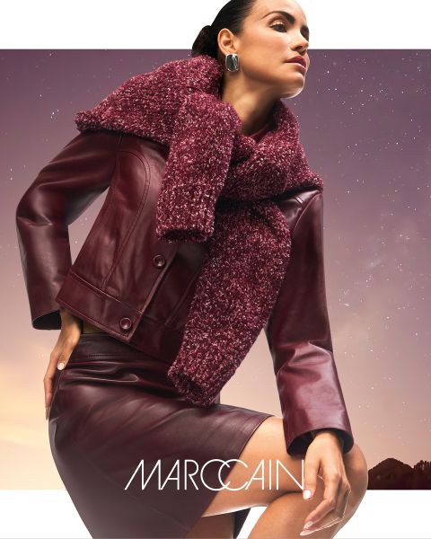 Una mujer posa con confianza con una chaqueta de cuero color burdeos y una falda con una bufanda gruesa y texturizada color burdeos. En el fondo se ve un cielo sombrío y el nombre de la marca "Marc Cain" se ve en la parte inferior.