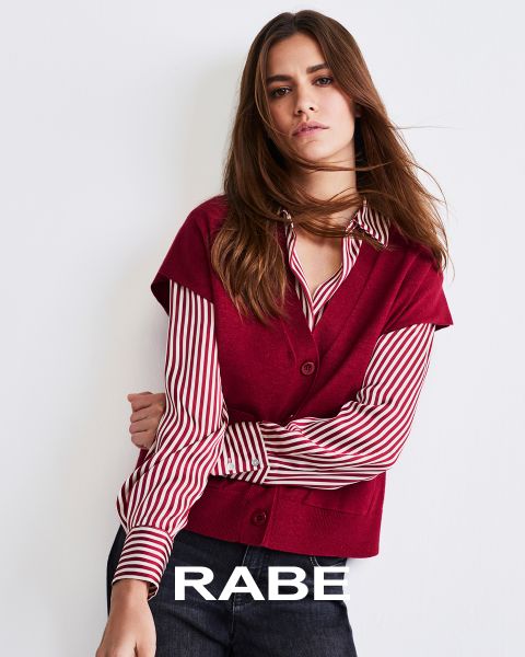Una mujer posa frente a una pared blanca, vistiendo un chaleco rojo sin mangas sobre una blusa a rayas rojas y blancas y pantalones oscuros. En la parte inferior de la imagen se puede leer la palabra "CUERVO".