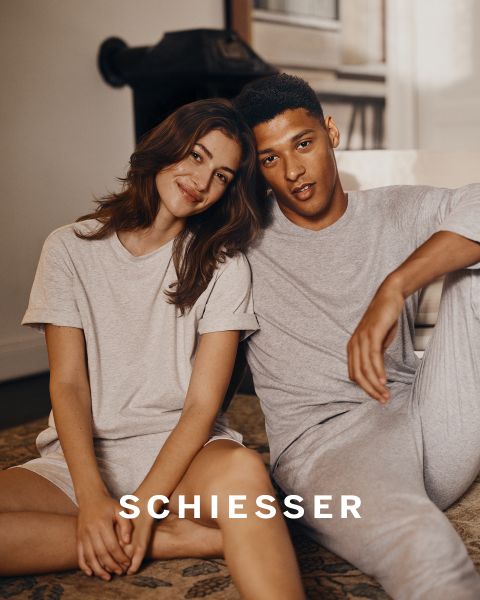 Una mujer y un hombre en ropa de estar por casa de color gris claro se sientan juntos en una alfombra y sonríen suavemente a la cámara. El nombre de la marca "SCHIESSER" se puede ver en la parte inferior de la imagen.