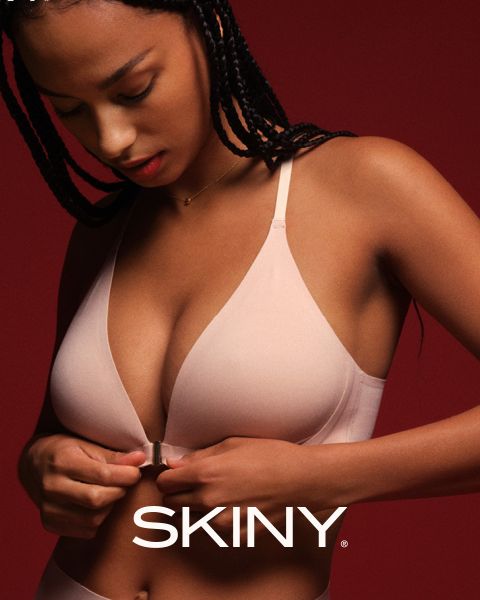 Una mujer con el pelo trenzado se abrocha un sujetador rosa claro con cierre frontal delante de un fondo rojo oscuro. La palabra "SKINY" aparece en la parte inferior de la imagen.