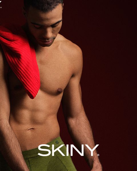 Un hombre musculoso y sencillo mira hacia abajo. Lleva un suéter rojo sobre el hombro y ropa interior verde. El fondo es de color rojo oscuro y la palabra "SKINY" aparece en la parte inferior.