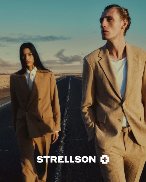 Un hombre y una mujer con elegantes trajes marrones caminan por una calle vacía al atardecer. El logotipo de Strellson aparece en la parte inferior de la imagen.