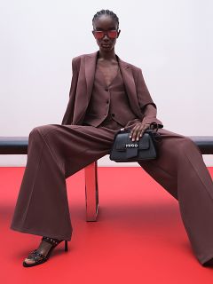 Una modelo con un traje marrón oversized, pantalones de pierna ancha y tacones negros está sentada en un banco frente a un fondo rojo y una pared blanca, lleva gafas de sol y sostiene un bolso negro de HUGO en la mano.