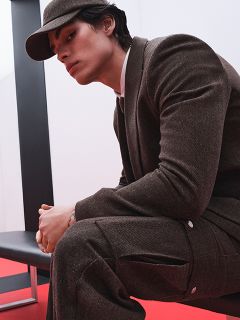 Una persona con un traje marrón y una gorra a juego está sentada en un banco negro frente a un fondo rojo y blanco, mirando hacia un lado con una expresión pensativa.