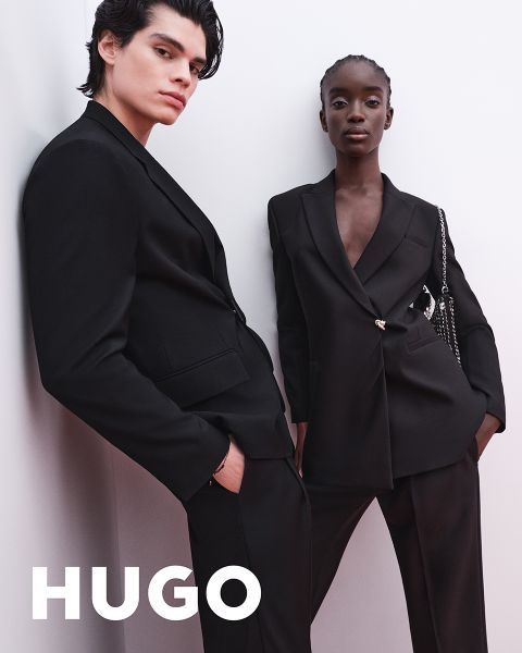 Dos modelos en trajes negros posan con confianza frente a un fondo claro y sencillo. El logo de HUGO aparece en letras blancas y gruesas en la esquina inferior izquierda.