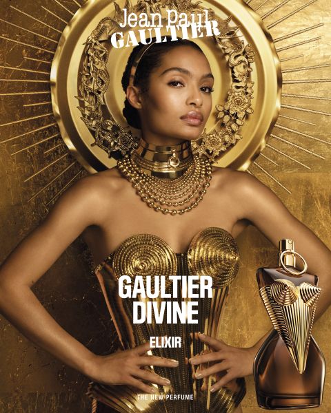 Una mujer con un vestido de metal dorado y joyas posa frente a un fondo dorado con efecto rayos de sol, con un frasco del perfume Jean Paul Gaultier Divine Elixir en primer plano. El nombre del perfume se muestra en negrita y en blanco.