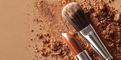 Una brocha de maquillaje y un lápiz labial color nude yacen sobre una superficie espolvoreada con polvos sueltos marrones, todos en tonos tierra similares.