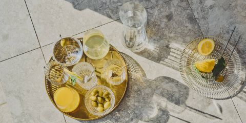 Una bandeja dorada con varias bebidas, aceitunas y un vaso de zumo de naranja está sobre una superficie alicatada a la luz del sol. Cerca hay un plato de cristal con rodajas de limón y una hoja, junto a una jarra de cristal con agua.