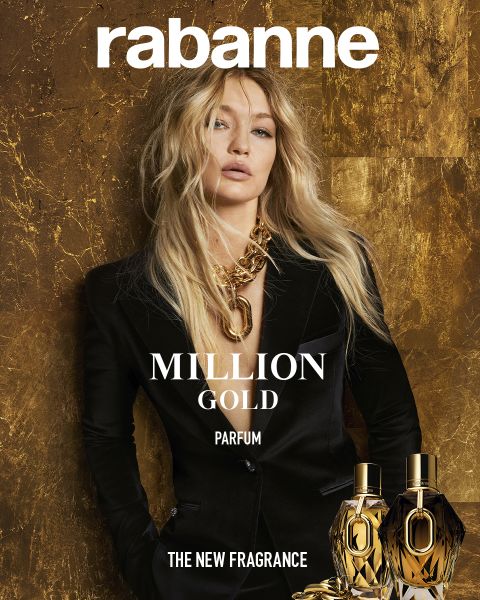 Un modelo con un traje negro y cabello rubio largo y ondulado lleva un collar dorado grueso y está de pie frente a un fondo con textura dorada. Arriba pone "rabanne", abajo pone "MILLION GOLD PARFUM" y dos frascos de perfume dorados.