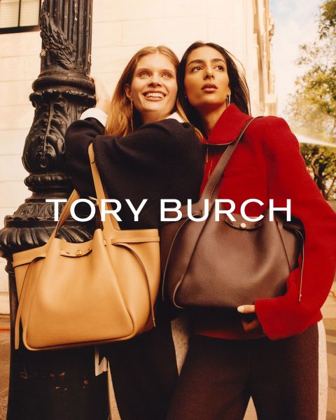 Dos mujeres están de pie al aire libre, cogidas del brazo, sonriendo, con abrigos elegantes y bolsos grandes de Tory Burch. La luz del sol resalta sus rostros. El texto "TORY BURCH" aparece sobre la imagen.
