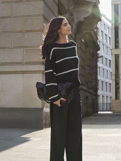 Una mujer con el pelo largo está de pie en una acera de la ciudad. Lleva un suéter negro con rayas blancas y pantalones negros de pierna ancha, y sostiene un bolso de mano negro grande mientras la luz del sol la ilumina desde atrás.