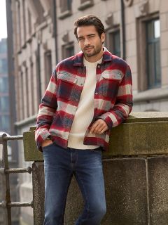 Un hombre con cabello corto y oscuro y barba está apoyado en una barandilla de piedra al aire libre. Lleva una chaqueta a cuadros roja y blanca sobre un suéter color crema y jeans azules. En el fondo se ven edificios urbanos.