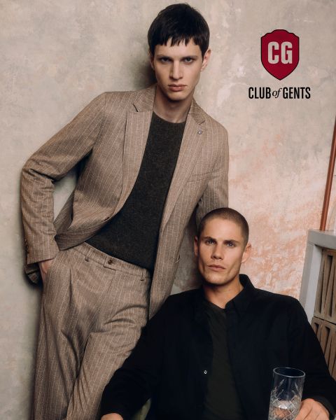Dos hombres posan para una foto de moda; uno lleva un traje de raya diplomática marrón claro y una camisa oscura, mientras que el otro está sentado a su lado con una camisa negra. El fondo está ligeramente texturizado, con el logo del "CG Club of Gents" encima de ellos.