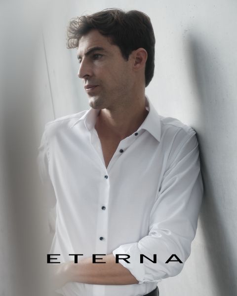 Un hombre con una camisa blanca se apoya en una pared clara y mira pensativo hacia un lado. En la parte inferior de la imagen está la palabra "ETERNA".