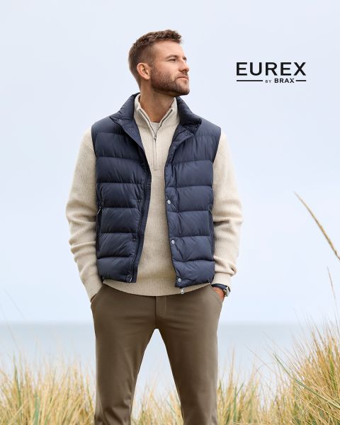 Un hombre está de pie al aire libre cerca de hierba alta, lleva un jersey beige, un chaleco acolchado azul marino y pantalones marrones, y mira hacia un lado. El logotipo de "EUREX by BRAX" aparece en el lado derecho. El cielo está nublado.