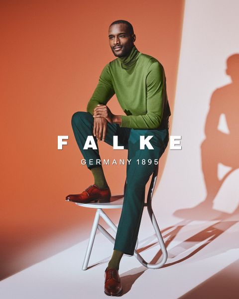 Un hombre con un jersey de cuello alto verde, pantalones verde oscuro y zapatos marrones está sentado en un taburete blanco frente a un fondo naranja y blanco. El texto "FALKE ALEMANIA 1895" está superpuesto a la imagen.