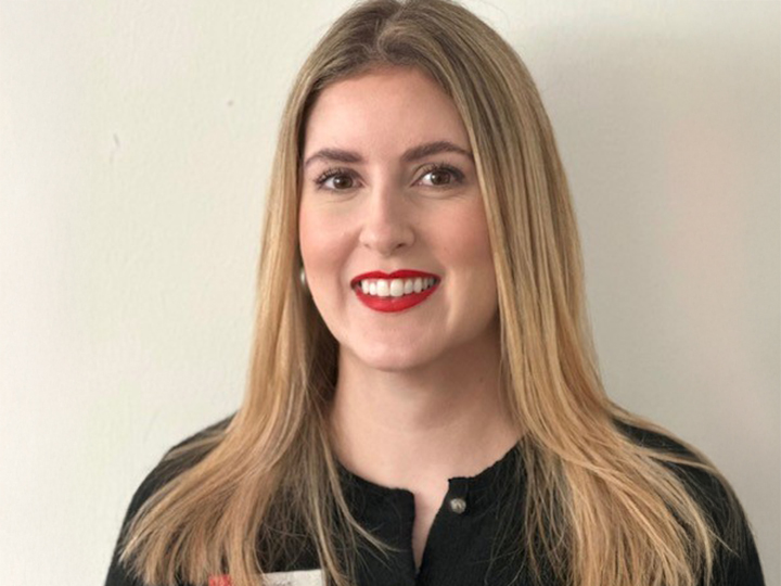 Mujer con cabello largo y rubio y lápiz labial rojo, que lleva una blusa negra, sonríe frente a un fondo claro y sencillo.