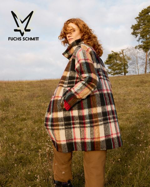 Una persona con cabello rizado, que lleva un abrigo ancho a cuadros y pantalones marrones, está de pie en un campo cubierto de hierba bajo un cielo nublado. En la esquina superior izquierda aparece el logo de Fuchs Schmitt.